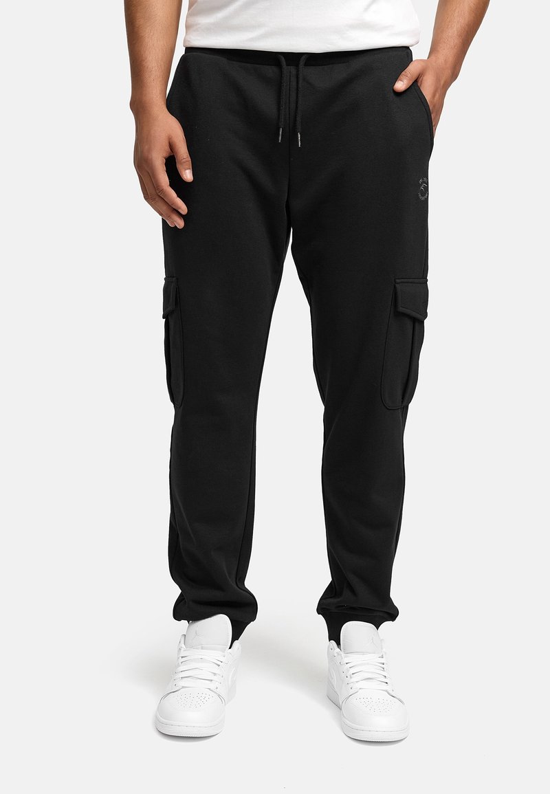 Homme portant un pantalon jogger cargo noir avec taille à cordon et des baskets blanches, debout devant un fond clair uni.