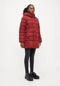 Rdeča puhovka z vodoravnim prešitim dizajnom, visokim ovratnikom in sprednjimi žepi. Ujemanja z črnimi oprijetimi kavbojkami in čevlji na vezalke.