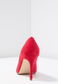 Czerwony but na wysokim obcasie wykonany z miękkiej tkaniny, z ostrym noskiem i smukłym obcasem. Wysokość obcasa wynosi około 10 cm. Gładka faktura.