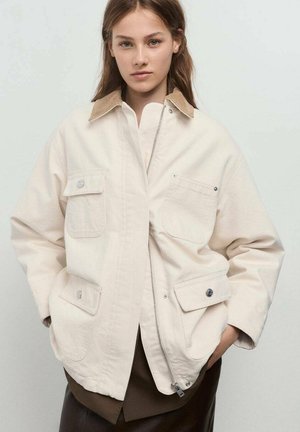 Beige canvas jacket with a tan collar, front zip closure, two large pockets, and silver hardware accents. Relaxed fit design.