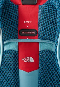 Mėlynas tinklinis audinys ir raudona medžiaga su tekstu "OPTI FIT". Yra reguliuojamas slankiklis, pažymėtas "177/43cm", ir "THE NORTH FACE" logotipas apačioje.