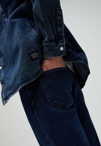 Jeansjacka i mörkblått med knappstängning, prydd med en patchetikett på ärmen. Matchad med mörkblå jeans.