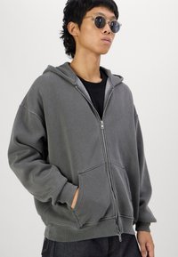 Grå zip-up hoodie med en mjuk textur, stora framfickor, justerbar huva och en avslappnad passform. Förstärkt med en svart t-shirt och runda solglasögon.