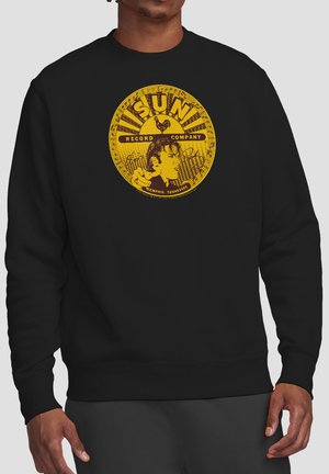Man wearing black sweatshirt with large yellow vintage Sun Record Company logo featuring a man’s profile and a rooster on the chest.