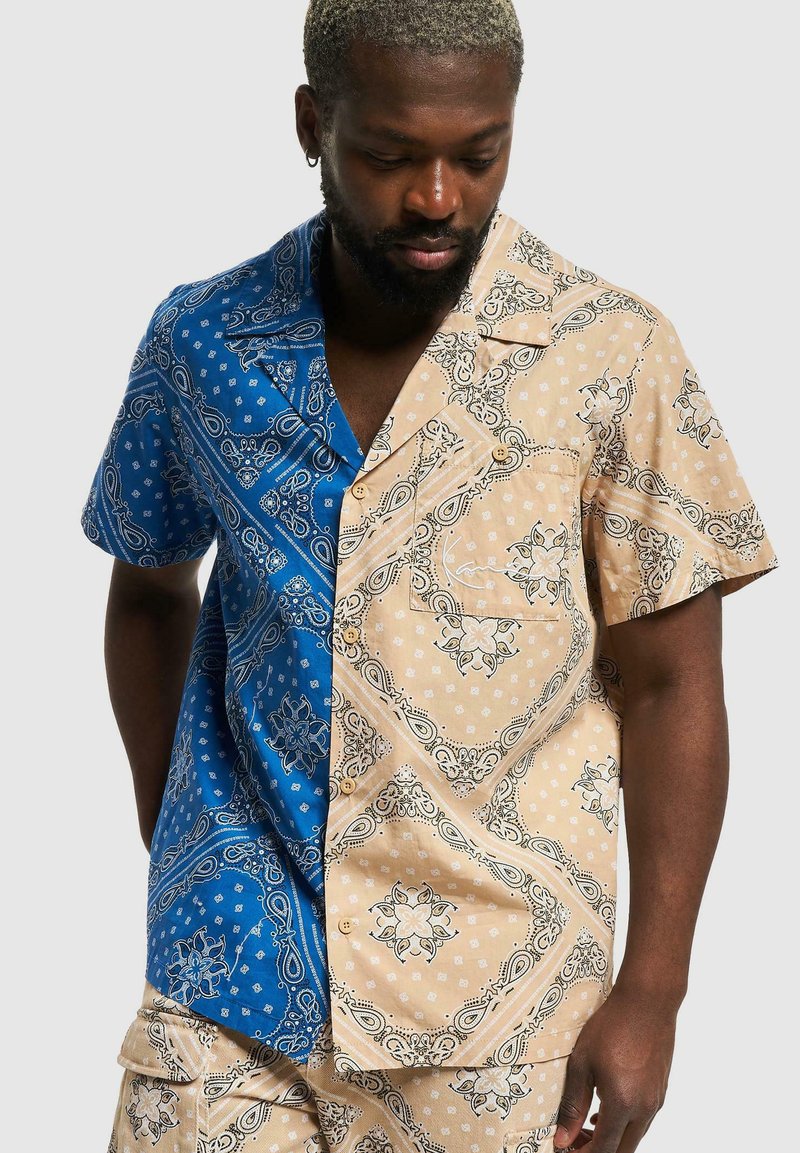 Kortermet skjorte med et blått og beige paisleymønster. Materialet er glatt, med en knappe-opp design og kontrasterende farger på hver side.
