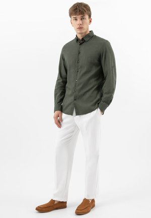 Giovane uomo in piedi, indossa una camicia verde scuro con bottoni, pantaloni bianchi e mocassini marroni, su uno sfondo bianco semplice.