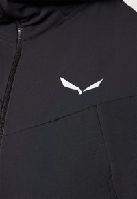 Veste noire en tissu lisse et extensible ; dotée d'une fermeture éclair complète et d'un logo blanc sur la poitrine ; design minimaliste avec une coupe ajustée.
