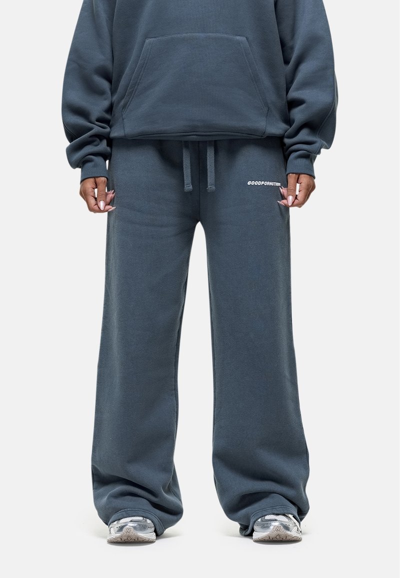 Haut à capuche oversized gris foncé avec une poche frontale et un pantalon de survêtement large assorti. Les deux pièces présentent un tissu doux et texturé ainsi qu'un logo imprimé.
