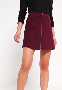 Burgundowa mini spódniczka w dopasowanym fasonie, z przednim zamkiem błyskawicznym i wyraźnie zarysowanymi szwami, uszyta z gładkiego materiału o delikatnym połysku.