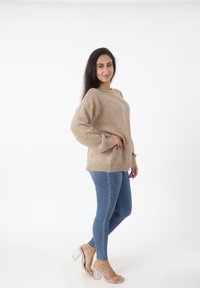 Beige gebreide trui met lange mouwen, geribde manchetten en zoom. G gedragen met blauwe skinny jeans en transparante hakken. Effen achtergrond.