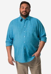 Světle modrá košile na knoflíky z jemné, texturované látky. Má jednu náprsní kapsu a ohrnuté rukávy. V kombinaci s béžovými kalhotami.