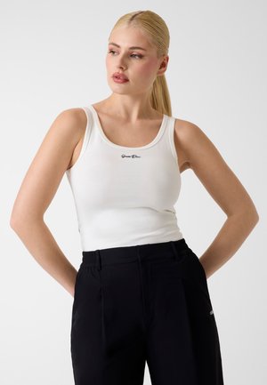 Femme aux longs cheveux blonds portant un haut blanc sans manches avec un texte noir et un pantalon noir, regardant vers la gauche avec les mains derrière le dos.