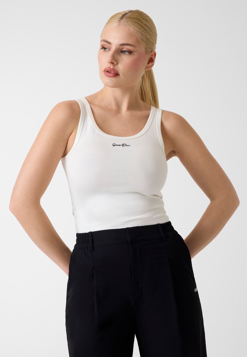 Femme aux longs cheveux blonds portant un haut blanc sans manches avec un texte noir et un pantalon noir, regardant vers la gauche avec les mains derrière le dos.