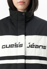 Nő fekete-fehér, magas nyakú dzsekit visel, amelyen fekete betűkkel hímzett "Guess Jeans" felirat található a mellkasán.
