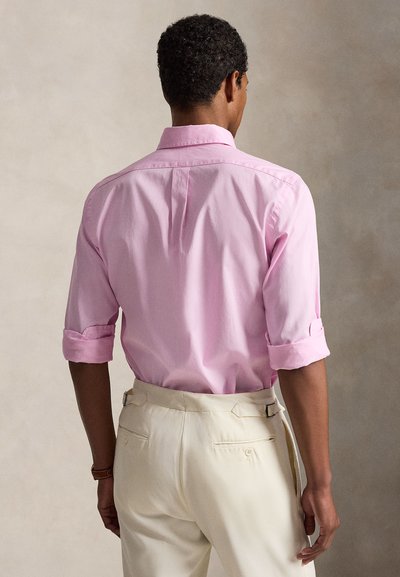 Άνδρας με μακριά μανίκια που έχει γυρίσει, φοράει ένα ανοιχτό ροζ πουκάμισο με κουμπιά που έχει βάλει μέσα σε ανοιχτό μπεζ παντελόνι, στέκεται μπροστά σε ουδέτερο φόντο.