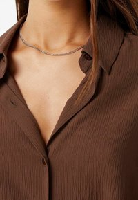Camicia marrone con texture, colletto e chiusura a bottoni. Collana in argento si posiziona all'altezza del collo, aggiungendo un dettaglio di accentuazione.
