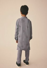 Ljust lila traditionell klädsel med blommönster, lång jacka, matchande byxor och svarta slip-on skor, sedd bakifrån.