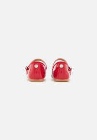 Chaussures en cuir verni rouge avec un talon bas et une sangle à boucle. Présentent une texture lisse et des accents métalliques ronds à l'arrière.