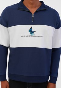 Nautična i bijela zip-up dukserica s dizajnom leptira i tekstom "KADA PROCVJETATE, LEPTIRI ĆE DOĆI K VAMA." Mekana, teksturirana tkanina.