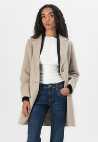 Beige Wollmantel mit tailliertem Design, Single-Button-Verschluss und Reverskragen, getragen über einer weißen Bluse und blauen Jeans.