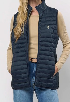 Kobieta ubrana w granatową pikowaną kamizelkę na beżowej bluzce z długim rękawem, niebieskie dżinsy i brązowy pasek, ręce w kieszeniach kamizelki.