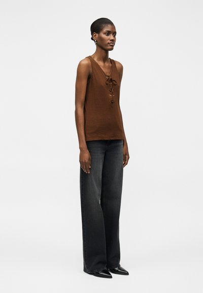 Γυναίκα με καφέ αμάνικη μπλούζα με κορδόνι μπροστά, μαύρο τζιν με φαρδιά μπατζάκια και μαύρα μυτερά παπούτσια, που στέκεται μπροστά σε απλό φόντο.