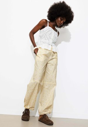 Femme aux cheveux afro naturels portant un haut blanc sans manches, un pantalon léger à fleurs avec cordon, des chaussures foncées sans lacets et un bracelet blanc, debout contre un mur blanc.