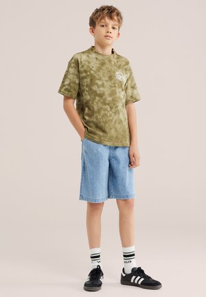 Dječak stoji sa rukama u džepovima, nosi zelenu majicu tie-dye, svijetloplave traper kratke hlače, bijele čarape i crne tenisice na jednobojnoj pozadini.