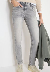 Jean en denim gris délavé avec une coupe slim, cinq poches et des détails de coutures visibles. Associé à un haut à manches longues et une veste verte.