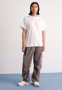 Mand med skulderlangt hår iført en hvid Nike t-shirt, grå cargo bukser og hvide sneakers, der står på et blåt gulv mod en hvid væg.