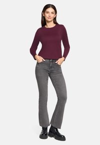 Bordeauxrode gebreide trui met ronde hals, gecombineerd met grijze strakke spijkerbroek en zwarte chunky enkellaarsjes, gepresenteerd tegen een effen achtergrond.