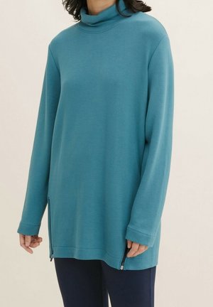 Frau trägt einen türkisfarbenen Rollkragenpullover mit langen Ärmeln und seitlichen Reißverschlüssen sowie eine dunkle Hose, steht vor einem einfarbigen hellen Hintergrund.