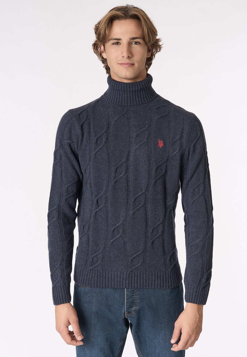 Giovane uomo che indossa un maglione a collo alto in maglia cable blu scuro e jeans blu, in piedi contro uno sfondo bianco semplice.