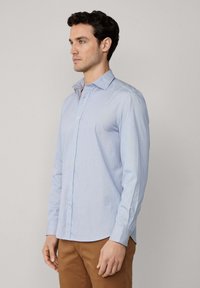 Camisa de botón azul claro con cuello puntiagudo, de manga larga y con sutiles rayas verticales, combinada con pantalones marrones.