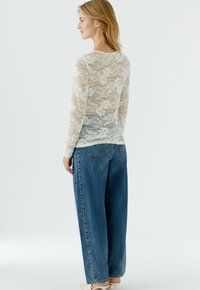 Lace long-sleeve top with floral pattern, cream color, paired with loose blue denim jeans. Back view shows sheer material and relaxed fit.