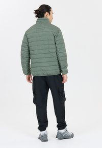 Polstret olivengrøn jakke med høj krave, præsenterer et horisontalt quiltet design. Kombineret med sorte cargo-bukser og grå sneakers.