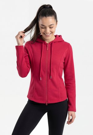 Junge Frau mit hohem Pferdeschwanz, die einen roten Kapuzenpullover mit Reißverschluss und schwarze Leggings trägt, lächelt, während sie eine Haarsträhne hält.