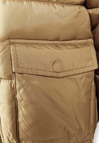 Jachetă puf din brown, în prim-plan, cu o mare buzunar cu închidere prin capsa, textură netedă și design matlasat orizontal.