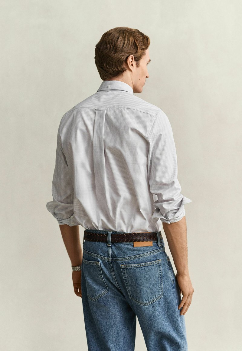 Mann mit weißem kariertem Langarmhemd mit hochgekrempelten Ärmeln und blauer Jeans, von hinten vor neutralem Hintergrund zu sehen.