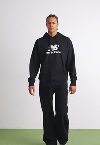 Fekete kapucnis pulóver fehér "New Balance" logóval, kenguru zsebbel és zsinórral. Fekete nadrággal viselve, zöld felületen állva.