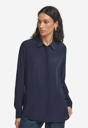 Kobieta o długich ciemnych włosach, ubrana w ciemnoniebieską bluzkę z długimi rękawami i kołnierzykiem, stoi na tle jednolitego białego tła.