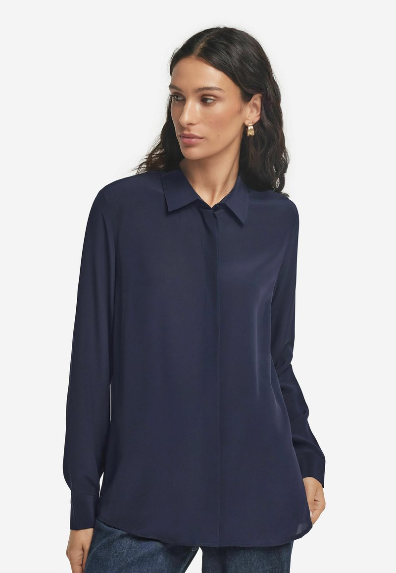 Kobieta o długich ciemnych włosach, ubrana w ciemnoniebieską bluzkę z długimi rękawami i kołnierzykiem, stoi na tle jednolitego białego tła.