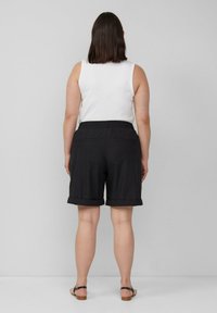 Schwarze Bermuda-Shorts mit umgeschlagenen Säumen, elastischem Bund und zwei Gesäßtaschen. Kombiniert mit einem weißen ärmellosen Oberteil und Sandalen.