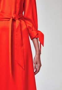 Robe rouge avec ceinture nouée à la taille et manchette assortie nouée sur la manche, montrant une partie du bras et de la main du modèle sur un fond uni.