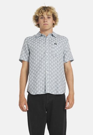 Jonge man met krullend blond haar, gekleed in een grijs kort mouwig overhemd met patroon en een zwarte broek, staand tegen een effen achtergrond.