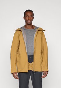 Żółta kurtka wodoodporna z szarym podkładem, kapturem i zapięciem na zamek. Posiada elastyczne mankiety i jest zestawiona z szarym ubraniem pod spodem.