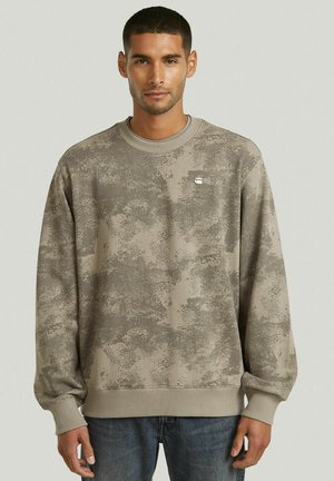 Mann stehend, trägt ein beiges und grau strukturiertes Langarm-Sweatshirt sowie blaue Jeans, blickt nach vorne mit neutralem Gesichtsausdruck.