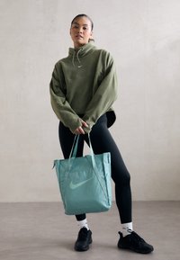 Vrouw in groene fleecetrekker, zwarte leggings, zwarte sneakers en witte sokken houdt een groene Nike draagtasje vast tegen een plain muren achtergrond.