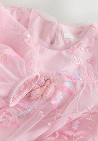 Vaaleanpunainen mekko, jossa on tylli ylle, esittäen brodeerattuja kukkakuoseja ja kimallekoristeita. Pyöreä pääntie ja puffimaiset hihat.