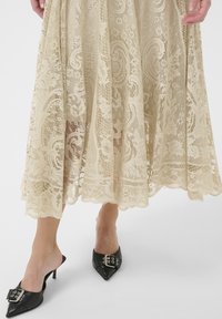 Gonna in pizzo beige con motivi floreali intricati, orlo scallop; abbinata a mule nere a punta con una fibbia decorativa.
